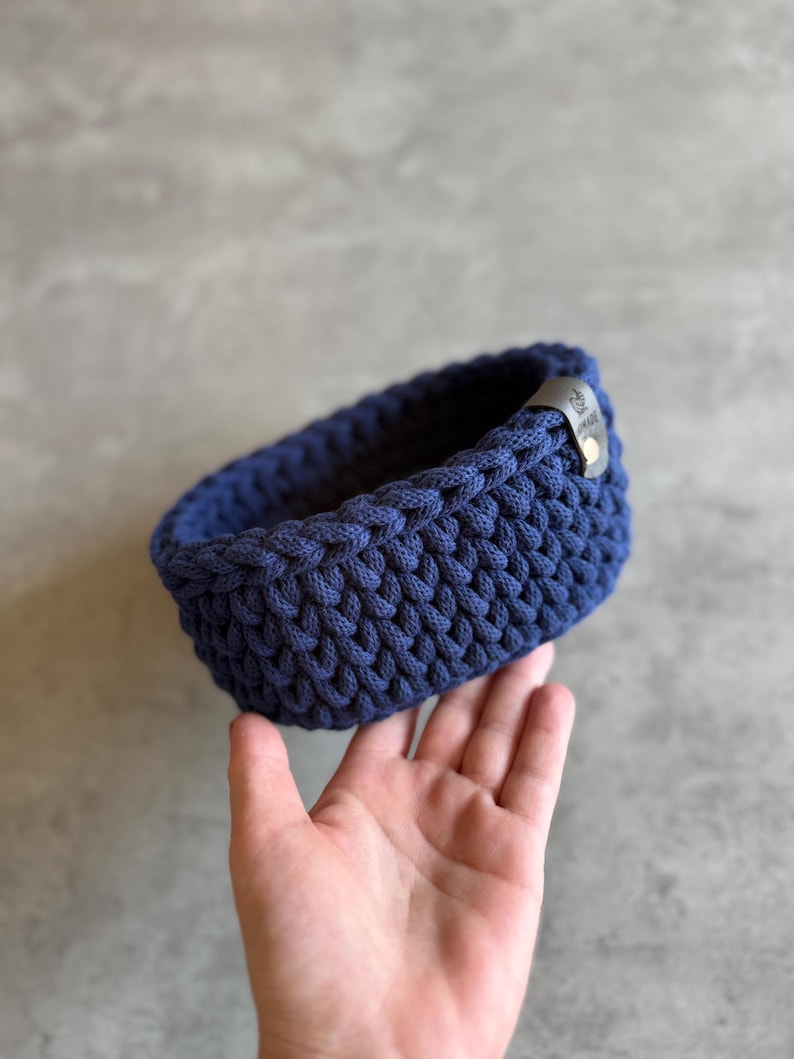 May include: A navy blue, hand-crocheted basket with a rounded rectangular shape. The basket is made of thick, textured yarn and features a small, dark leather tag. The basket is held in a hand against a gray background.