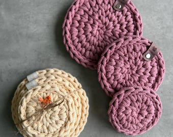 May include: Four handmade crochet coasters in shades of pink and cream. The pink coasters are in varying sizes, with a leather tag attached. The cream-colored coasters are bundled together with twine and a small dried flower.