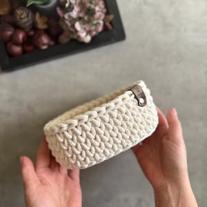 May include: A cream-colored, rectangular crochet basket with a leather tag. The basket is held in two hands. The basket is made of thick, textured yarn. The background includes a decorative tray with plants and dried flowers.