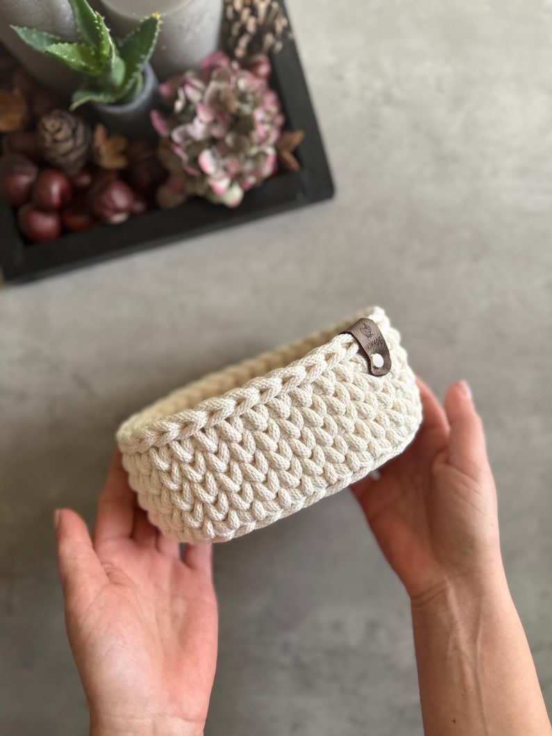 May include: A cream-colored, rectangular crochet basket with a leather tag. The basket is held in two hands. The basket is made of thick, textured yarn. The background includes a decorative tray with plants and dried flowers.