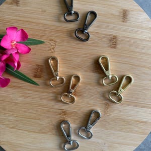 Könnte beinhalten: Eine Auswahl an Metall-Drehhaken in den Farbtönen Gunmetal, Gold und Silber. Die Haken sind auf einer runden Holzoberfläche angeordnet, mit einer rosa Blume und grünen Blättern. Ideal für Schlüsselanhänger, Schlüsselbänder und Bastelprojekte.