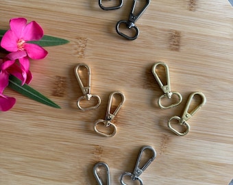 May include: An assortment of metal swivel clasps in gunmetal, gold, and silver tones. The clasps are arranged on a round wooden surface, with a pink flower and green leaves in the corner. These clasps are ideal for keychains, lanyards, and other craft projects.