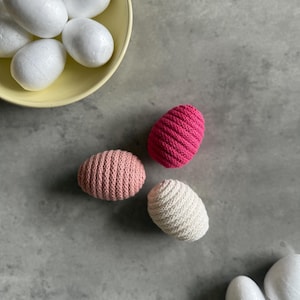 Könnte beinhalten: Draufsicht auf eine gelbe Schüssel mit weißen Eiern und drei farbigen, gehäkelten Eiern. Die gehäkelten Eier sind rosa, hellrosa und weiß. Der Hintergrund ist eine graue Oberfläche.