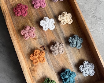 May include: A wooden tray holds an assortment of crocheted flower appliques in various colors. The flowers are arranged in a grid pattern, showcasing shades of pink, white, orange, green, blue, and neutral tones. The tray's natural wood grain provides a warm contrast.