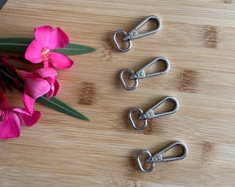 May include: Four silver-colored swivel clasps with a spring-loaded gate are arranged on a round wooden cutting board. Pink flowers and green leaves are in the upper left corner. The clasps are commonly used for keychains and lanyards.