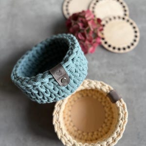 Könnte beinhalten: Zwei handgefertigte Häkelkörbe, einer hellblau und einer cremefarben, mit Lederetiketten. Die Körbe sind rund und haben ein strukturiertes Stichmuster. Untersetzer aus Holz und rosa Blumen sind im Hintergrund.