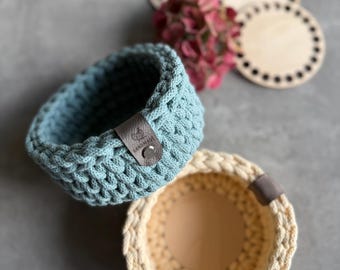 May include: Two handmade crochet baskets, one light blue and one cream, with leather tags. The baskets are round and have a textured stitch pattern. Wooden coasters and pink flowers are in the background.