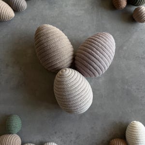 Könnte beinhalten: Gestrickte Eier-Dekorationen in verschiedenen neutralen Farben wie Beige, Creme und Olivgrün. Die Eier haben eine gerippte Textur und sind auf einer grauen Oberfläche angeordnet. Ideal für Oster- oder Frühlingsdekorationen.