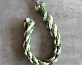 May include: A braided keychain wrist strap in green and white. The strap is made of thick, woven rope and features a silver-colored metal clasp. The design incorporates a repeating pattern of green and white strands.