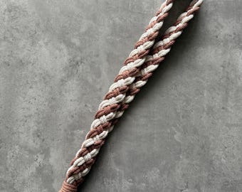 May include: Braided lanyard in a macrame style, featuring alternating strands of white and muted rose-colored cord. The lanyard has a loop at the top and a gold-colored metal clasp at the bottom for attaching keys or an ID badge.