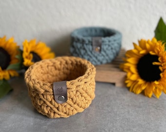 May include: Two crocheted baskets, one mustard yellow and one light blue, with leather handles. The baskets are sitting on a wooden surface with sunflowers in the background.