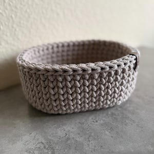 May include: A small, oval-shaped, light gray crochet basket. The basket is made of thick yarn, with a textured stitch pattern. The rim is finished with a braided edge. The basket is suitable for storing small items.