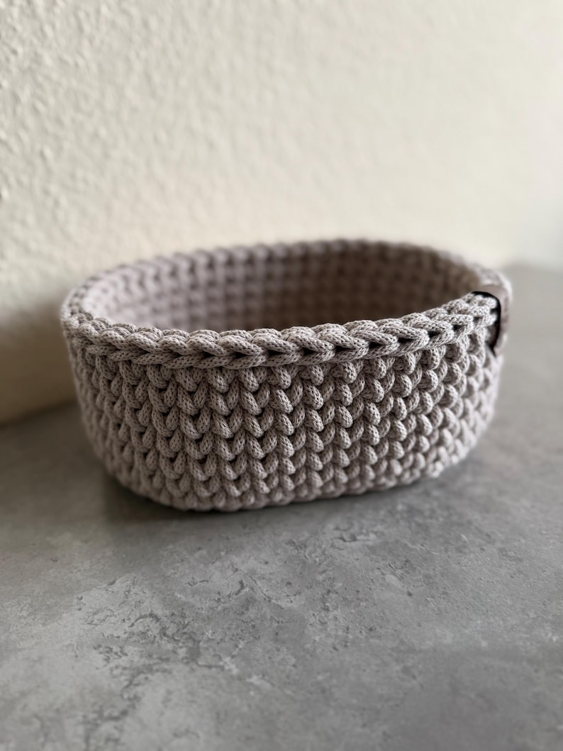 May include: A small, oval-shaped, light gray crochet basket. The basket is made of thick yarn, with a textured stitch pattern. The rim is finished with a braided edge. The basket is suitable for storing small items.