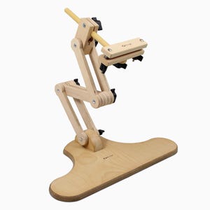 May include: A light-colored wooden embroidery stand featuring an adjustable articulated arm with multiple joints, black tightening knobs, and a clamp mechanism at the top. The base is T-shaped, and the brand name Ollurge is visible on the wood.