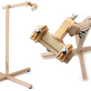 May include: A light-colored wooden stitching pony featuring a vertical stand with a cross-shaped base and an adjustable clamp head. The clamp mechanism includes metal wing nuts and silver-toned hardware to secure leather pieces during hand stitching.