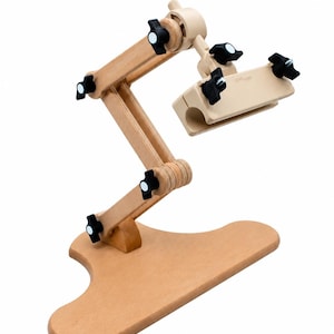 May include: A light-colored wooden adjustable stand featuring a flat base, a multi-jointed articulated arm, and a clamp mechanism at the top. The structure is held together by several black plastic knobs with white circular centers.