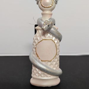 May include: A pale pink decorative bottle featuring a textured diamond pattern on the body and a smooth, circular blank label outlined in gold. A metallic silver snake is coiled around the neck and body, topped with a round, pearl-like stopper.