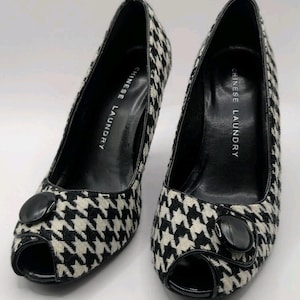 May include: A pair of black and white houndstooth peep-toe pumps with a high heel. Each shoe features a large, round, black decorative button on the outer side near the toe. The interior insole is black and printed with the text CHINESE LAUNDRY.