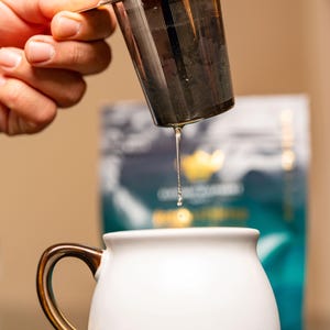 May include: A stainless steel tea infuser with a handle is being held over a white ceramic mug with a gold handle. A single drop of tea is falling from the infuser into the mug.