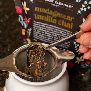 Puede incluir: Una tetera de cerámica blanca con un infusor de té de acero inoxidable. Se vierte té de hojas sueltas en el infusor desde una bolsa etiquetada "The Elegant Elephant Tea Co." con el texto "Madagascar Vanilla Chai".