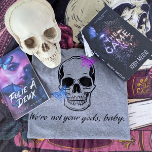 May include: A gray t-shirt with a black skull graphic and the text "We're not your gods, baby."  The shirt is displayed on a purple and black patterned fabric with two books, one titled "Folie a Deux" and the other titled "The Game".