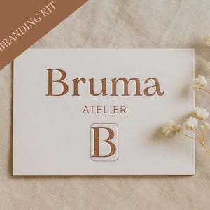 May include: A cream-colored business card with the name "Bruma" in embossed brown lettering, along with "ATELIER" and a stylized "B" logo. The words "MINI BRANDING KIT" are in a brown banner in the upper left corner. White flowers are in the background.