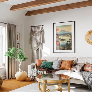 Könnte beinhalten: Ein gemütliches Wohnzimmer mit einem beigen Sofa, das mit bunten Kissen und einer Kunstfelldecke geschmückt ist. Über dem Sofa hängt ein gerahmtes Landschaftskunstwerk. Der Raum verfügt über einen runden Couchtisch, einen geflochtenen Hocker und einen kleinen Baum in einem großen Topf.