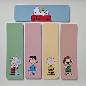 May include: A set of five rectangular bookmarks featuring Peanuts characters. One horizontal bookmark shows Snoopy and Woodstock on a red doghouse. Four vertical bookmarks in green, pink, yellow, and tan feature Linus, Lucy, Charlie Brown, and Peppermint Patty.