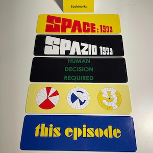 May include: A set of six bookmarks featuring the title "Space: 1999" in various languages and designs. The bookmarks are yellow, black, and blue with white and red text. One bookmark features a circular design with numbers and a red, white, and blue colour scheme.