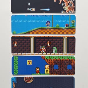 May include: A set of five rectangular coasters featuring pixelated video game scenes. The designs include a space shooter, a blue hedgehog on a grassy hill, a knight in a stone corridor, a character in a platformer level, and a dark dungeon scene.