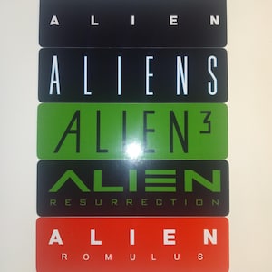 May include: A stack of five black, white, green, and red stickers with the words "Alien", "Aliens", "Alien 3", "Alien Resurrection", and "Alien Romulus" printed on them.