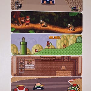 May include: Rectangular stickers featuring scenes from classic video games. The top sticker shows a racing game with a track and vehicles. Other stickers depict scenes from Donkey Kong, Super Mario, and The Legend of Zelda.