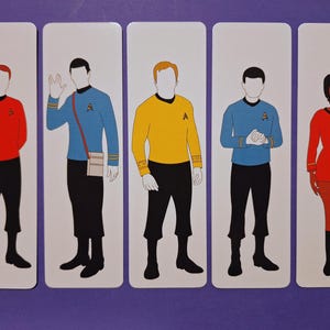 Op de afbeelding: Vijf rechthoekige kaarten met gestileerde illustraties van Star Trek-personages. Elke figuur draagt een uniform in een andere kleur: rood, blauw en geel. De figuren staan tegen een witte achtergrond.