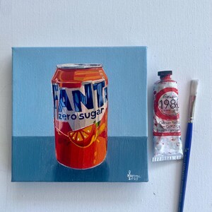 May include: A painting of a Fanta Zero Sugar can with an orange slice illustration. A tube of red paint and a paintbrush are next to the can. The background is light blue and dark blue.