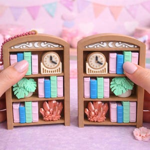 May include: Two miniature wooden bookshelves, each with a clock, books in pastel colors, a plant, and a crystal. The shelves are held by hands against a soft pink background, suggesting a whimsical theme.