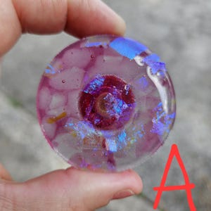 May include: A round, clear glass art piece with a central dark red design surrounded by pink and iridescent blue accents. The glass has a textured appearance, with a small gold-colored element in the center.