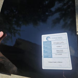 May include: A square sheet of black solid opalescent glass with a glossy, reflective surface. A white rectangular label is affixed to the bottom right corner, featuring the Oceanside Glass & Tile logo, product code 1009S-F, and production date 6/13/2025.