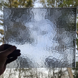 May include: A rectangular piece of textured glass held against a blurred outdoor background. The glass has a rippled surface, creating a distorted view. The edges of the glass are visible, and the overall color is a translucent gray.