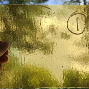 May include: A rectangular sheet of translucent yellow stained glass with a textured, rippled surface. The glass features a handwritten number one enclosed in a circle in the upper right corner, visible through the semi-transparent material.