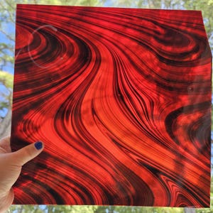 May include: A square sheet of translucent stained glass featuring a vibrant red base color with swirling, marbled patterns of deep black streaks throughout the surface. The glass has a smooth, polished texture and allows light to pass through it.