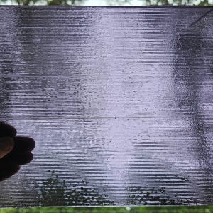 May include: A rectangular, translucent panel with a textured, linear pattern. The panel is held by a hand, and the background is blurred, suggesting an outdoor setting. The panel has a purplish hue.