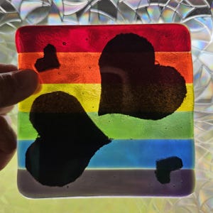 May include: A square glass coaster featuring horizontal stripes in red, orange, yellow, green, blue, and purple. The surface is decorated with four solid black heart shapes of varying sizes, arranged across the rainbow-colored background.