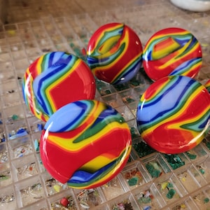 May include: A collection of five colorful, round glass marbles. Each marble features a vibrant swirl of red, yellow, blue, and green, creating a rainbow effect. The marbles are displayed on a grid-like surface, showcasing their unique patterns.