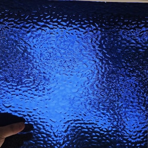 May include: A square piece of textured, cobalt blue glass. The glass has a rippled surface, creating a dynamic play of light and shadow. The color is a deep, saturated blue, with a slight variation in tone across the surface.