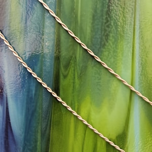 May include: A textured sheet of stained glass featuring a marbled blend of deep blue, emerald green, and lime green hues. Two thin, light-colored twisted metal wires run diagonally across the surface of the glass, creating a contrasting linear pattern.