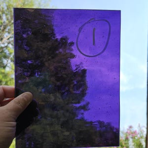 May include: A rectangular sheet of translucent purple glass with a textured surface containing small air bubbles. The number one is handwritten in a circle on the upper right portion of the glass surface, which is held up against a bright outdoor sky.