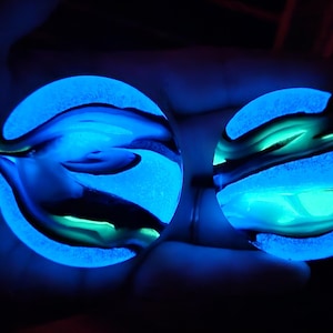 May include: Two glowing, round, blue-toned objects with swirling patterns of blue, green, and black. The objects appear to be illuminated from within, creating a vibrant, eye-catching effect. The background is dark, emphasizing the glowing objects.