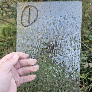 May include: A rectangular sheet of clear, textured glass featuring an irregular, hammered surface pattern. The upper left corner displays a hand-drawn circle containing the number one, marked in a dark, brownish-black ink.