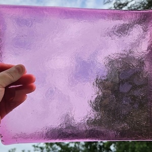 May include: A rectangular, translucent, pink glass plate with rounded corners. The glass has a textured surface and is held up against a sky background. The plate's color shifts from a lighter pink to a deeper shade, creating a gradient effect.