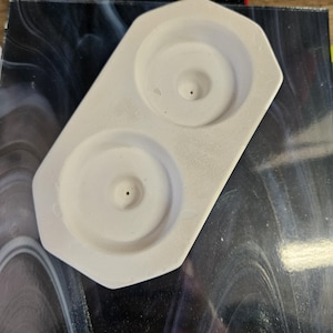 May include: A white, ceramic, two-well paint palette with a hexagonal shape. Each well has a central raised area with a small hole. The palette is designed for mixing and holding paints, inks, or other art mediums.